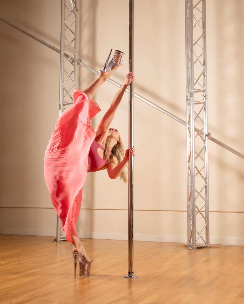 Jen M Pole Academy wearing a pink skirt and sports bra, lifting her leg high up wearing heels and holding a pole